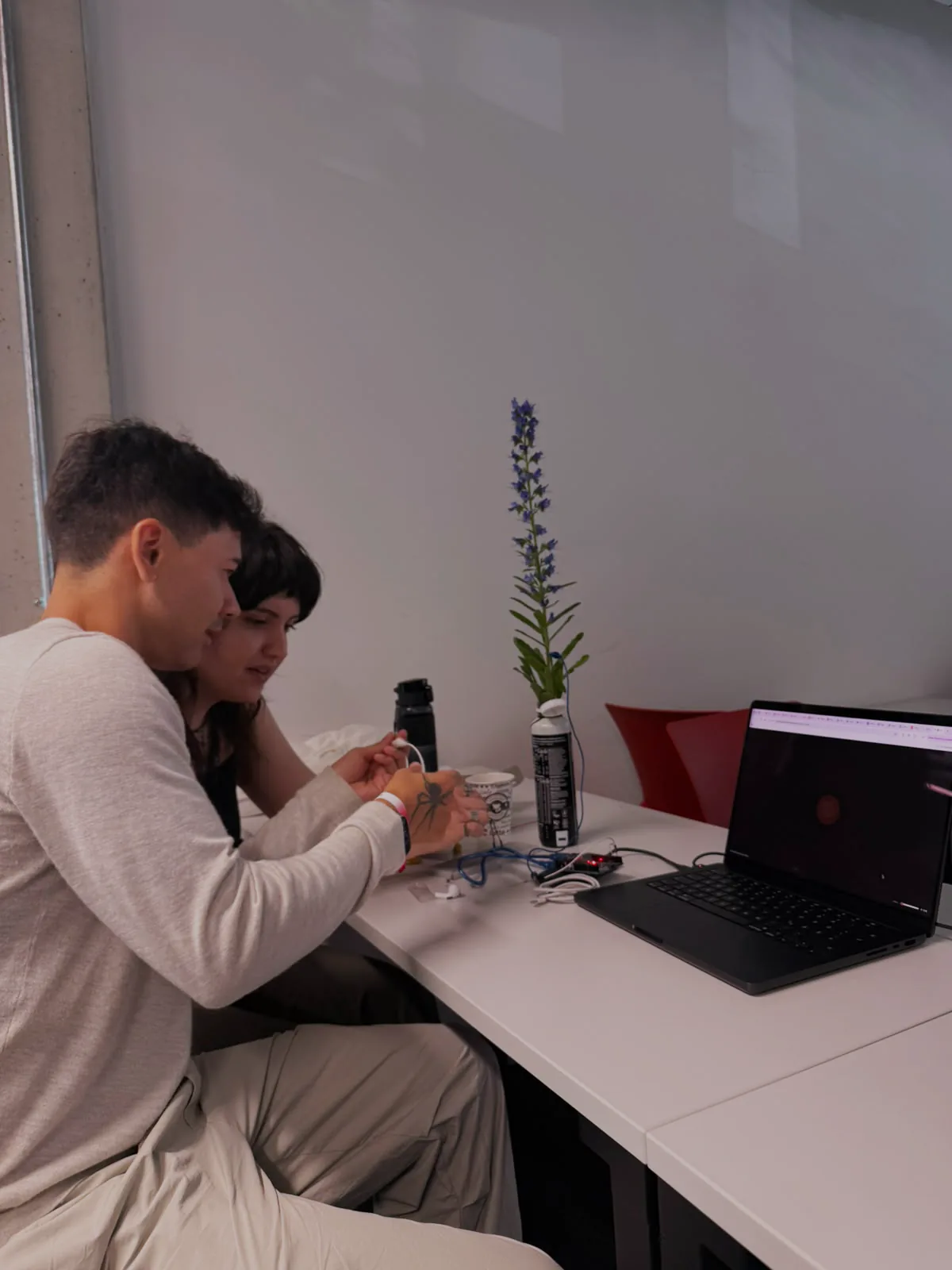 Ronald and Esra at a classroom desk, calibrating the piece: a flowering stem stands in a bottle wired to an Arduino, and both look at the laptop, where the particle sketch shows a single glowing point at rest.
